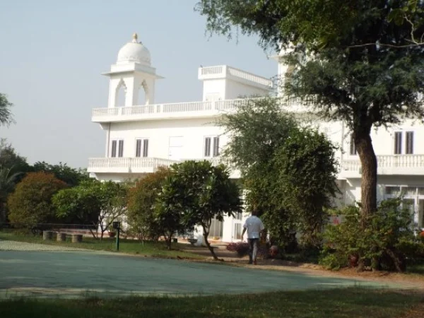 The image shows a sprawling white palace set within a vibrant green garden. The building features a central dome and multiple floors with prominent balconies. The architecture is classic Indian, with clean white lines and arched windows. In the foreground, various trees and bushes frame the scene, leading the eye towards the palace. A man is walking to the front door. The day is bright and sunny, casting shadows that give depth to the scene. The ground looks well-maintained with the lush grass and trees.
