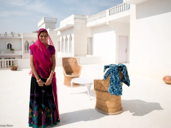 A medium shot of a woman in traditional Rajasthani clothing standing on a white terrace at the Savista Retreat. She is wearing a bright pink headcloth and a long, patterned skirt. Behind her are chairs and a small table.