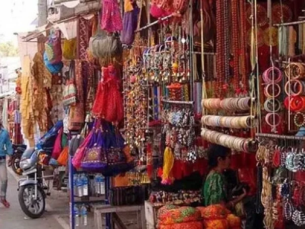 A bustling street market scene in Rajasthan, showcasing a vendor's stall filled with vibrant textiles, beaded jewelry, hanging fabrics, and stacks of colorful bangles.