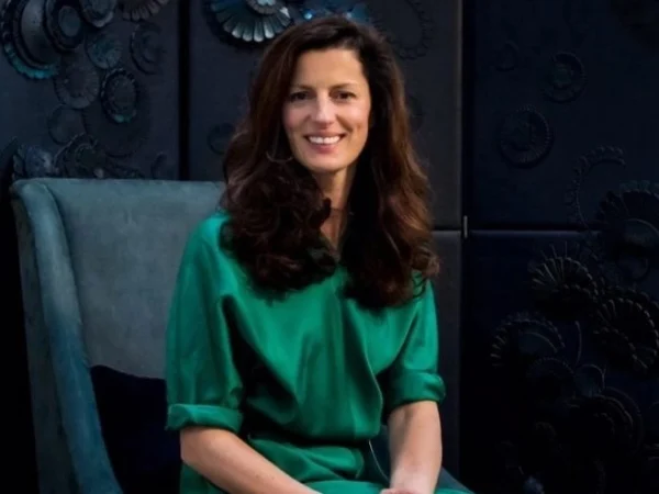 A medium shot of a woman seated in a teal-colored armchair. She has shoulder-length brown hair, and is wearing a bright green dress.