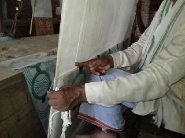 Close-up shot of a man with brown skin, wearing long-sleeved white and a blue-white checkered cloth around neck, sitting at a loom, the shuttle in his right hand. His left hand holds the yarn as he weaves.