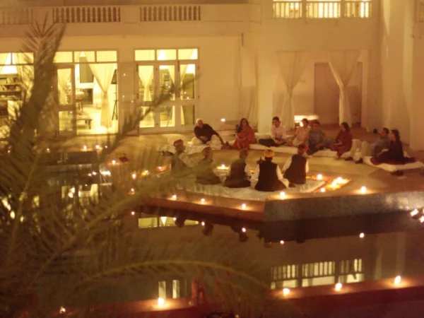 This indoor image captures the essence of Savista Retreat at night. A group of about 15 people is gathered around a rectangular reflective pool. The pool is illuminated by a ring of glowing candles, which casts reflections on the water.