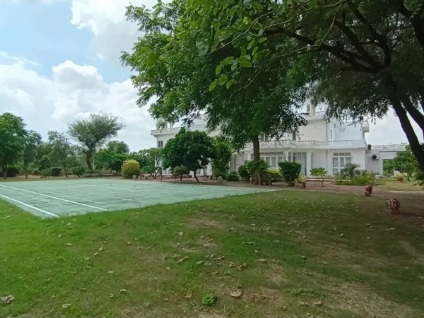 Savista Retreat: Tennis court, green lawn, and white building under a blue sky in Jaipur.