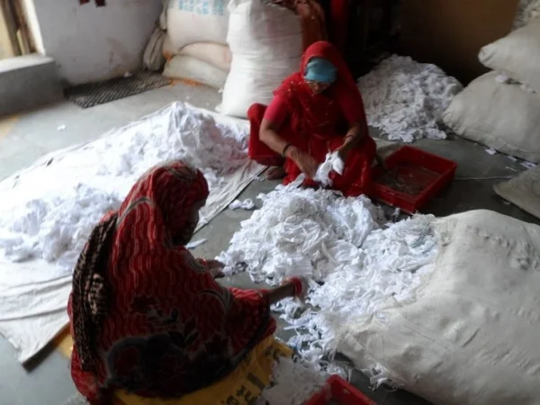 The image shows two women seated on the floor, sorting raw cotton. They are wearing traditional Indian clothing, including head coverings.