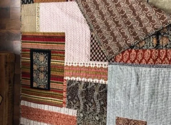 Textile patchwork quilt with assorted patterns and textures, partially overlaid by a beige rug featuring a decorative border.