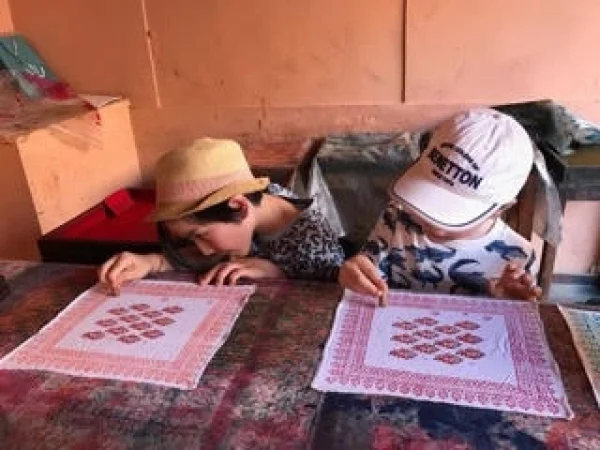 Two children, one wearing a hat and the other wearing a cap, concentrating on block printing designs on fabric in a Savista's workshop