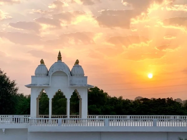 A savista retreat displays a serene sunset at its majestic white temple, three domes glistening under a golden roof. The bridge and tranquil setting create a haven of beauty, where warm hues blend with the retreats tranquil charm.